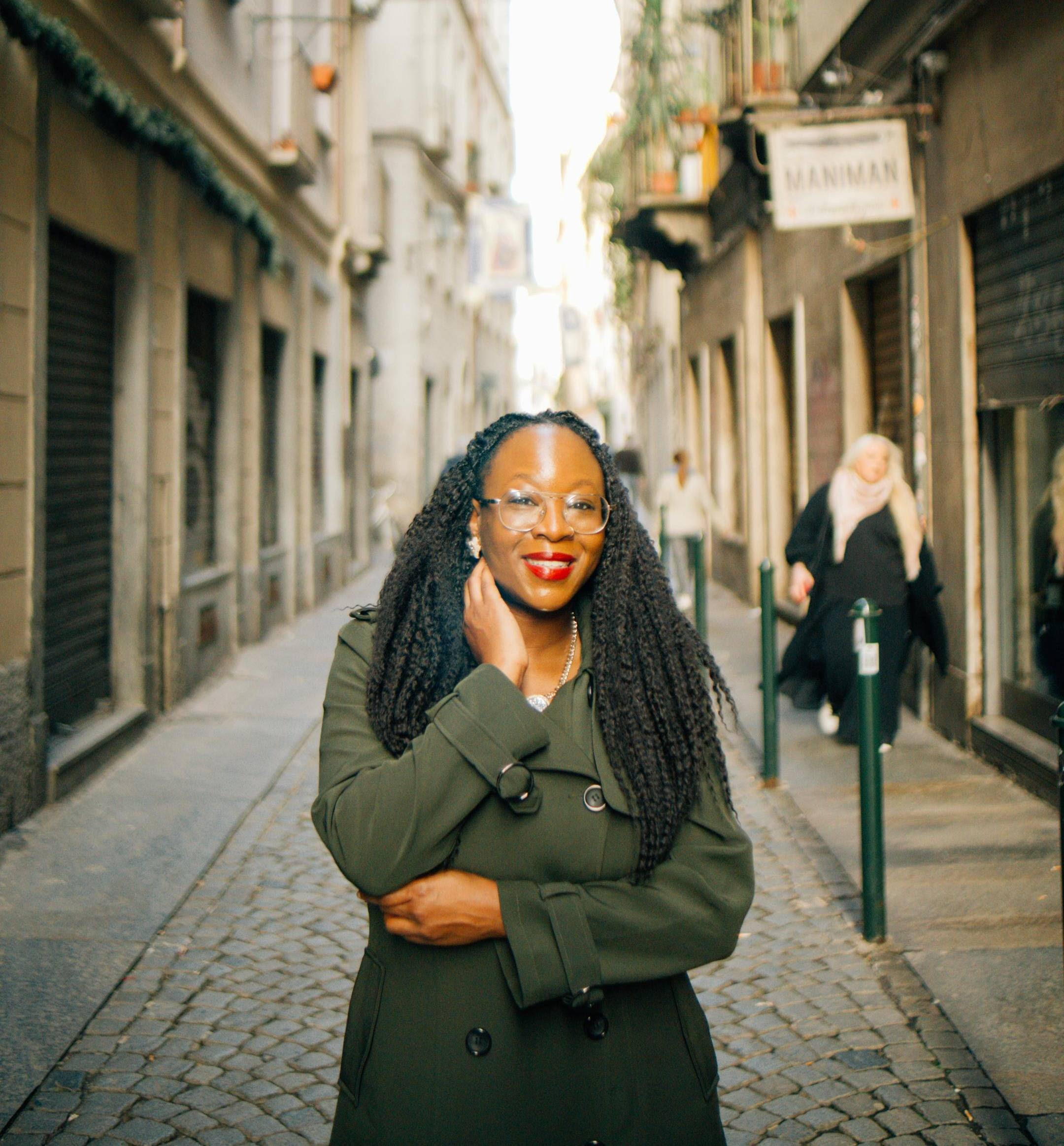 Portrait of Daphne, a life coach and founder of 'My Inner Fort,' standing confidently on a charming cobblestone street. With a radiant smile, stylish glasses, and a chic green coat, she embodies creativity, resilience, and adaptability in her approach to coaching. Ideal for anyone seeking guidance on personal growth, psychological strength, and embracing life’s journey.