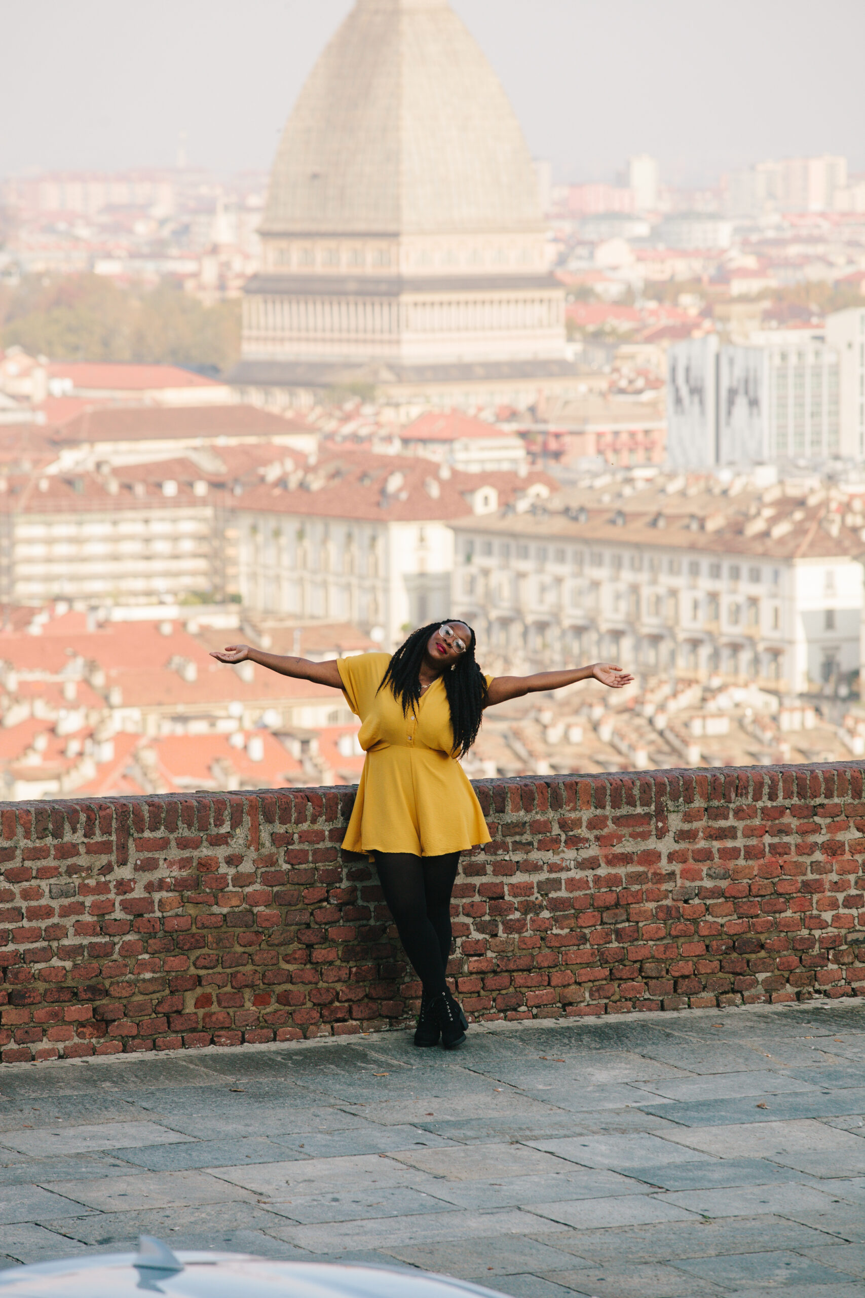 A joyful woman in a bright yellow dress standing against a brick wall with arms outstretched, overlooking a picturesque cityscape featuring iconic architecture and rooftops. The image exudes a sense of freedom, self-discovery, and the transformative power of travel.